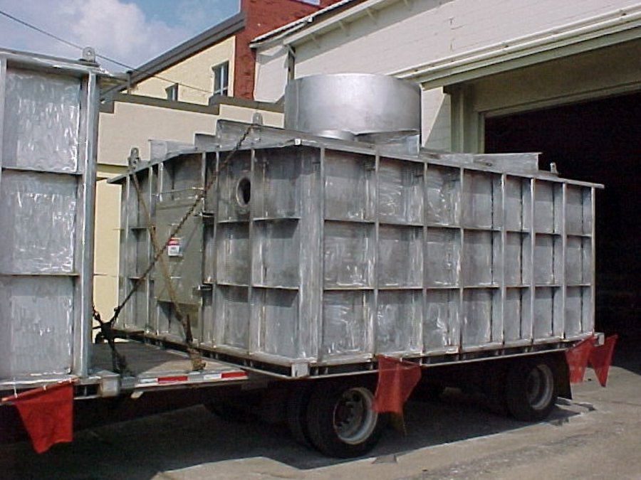design-build002 A large, custom-fabricated stainless steel industrial unit with multiple access ports and a top-mounted cylindrical stack is staged on a flatbed transport trailer.