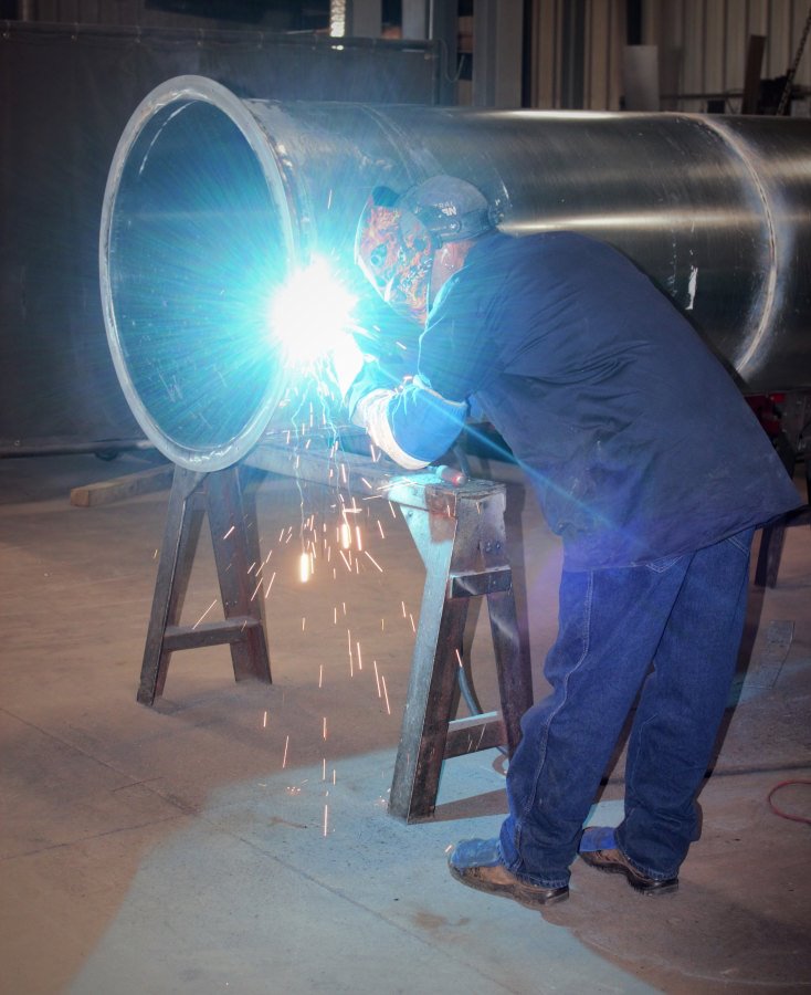 ductwork001 A technician in a protective welding helmet and blue workwear welds a large cylindrical metal pipe on a sawhorse, producing a bright blue arc and falling sparks.